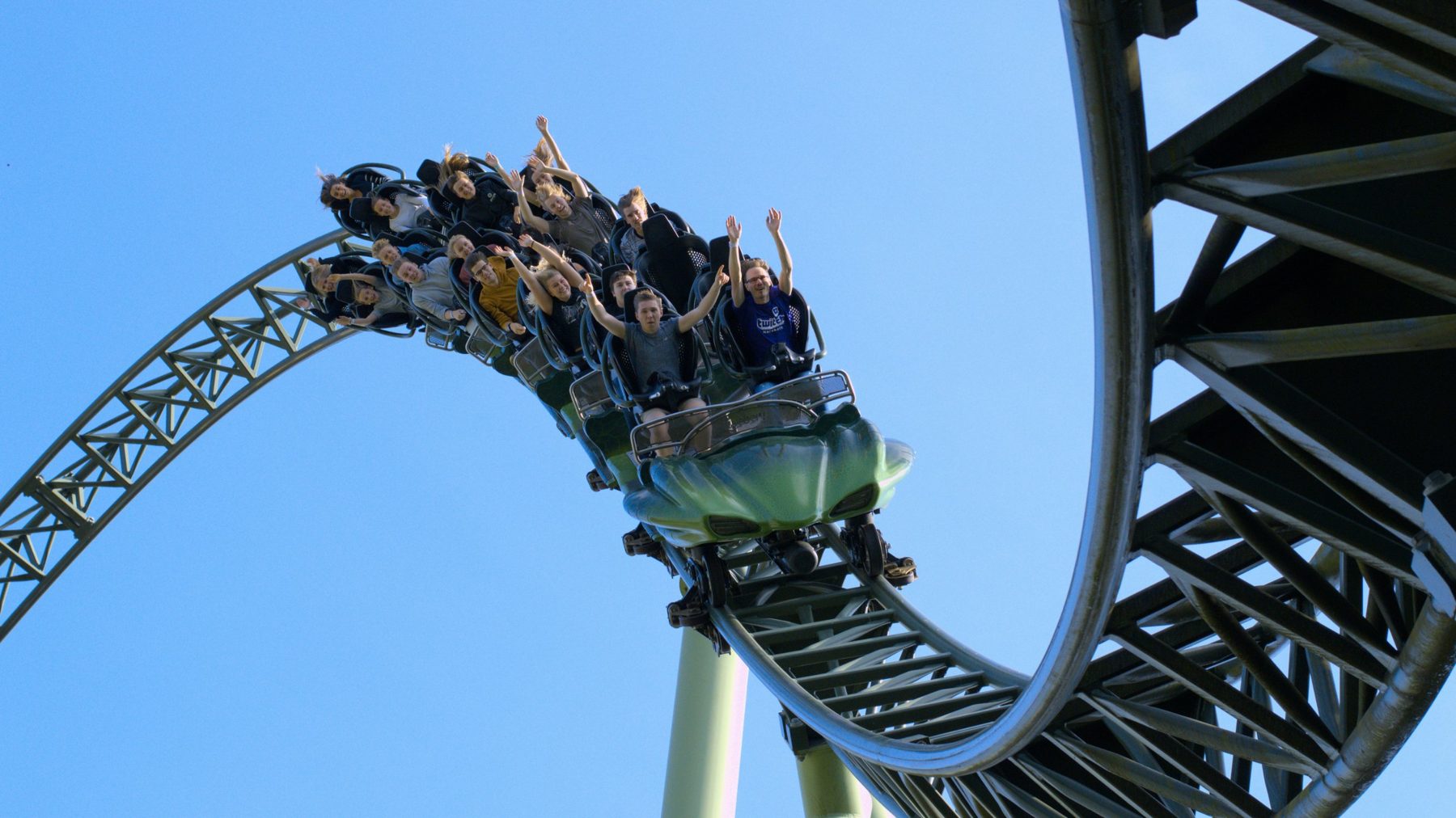 Riders with their arms in the air on a green roller coaster against a clear blue sky